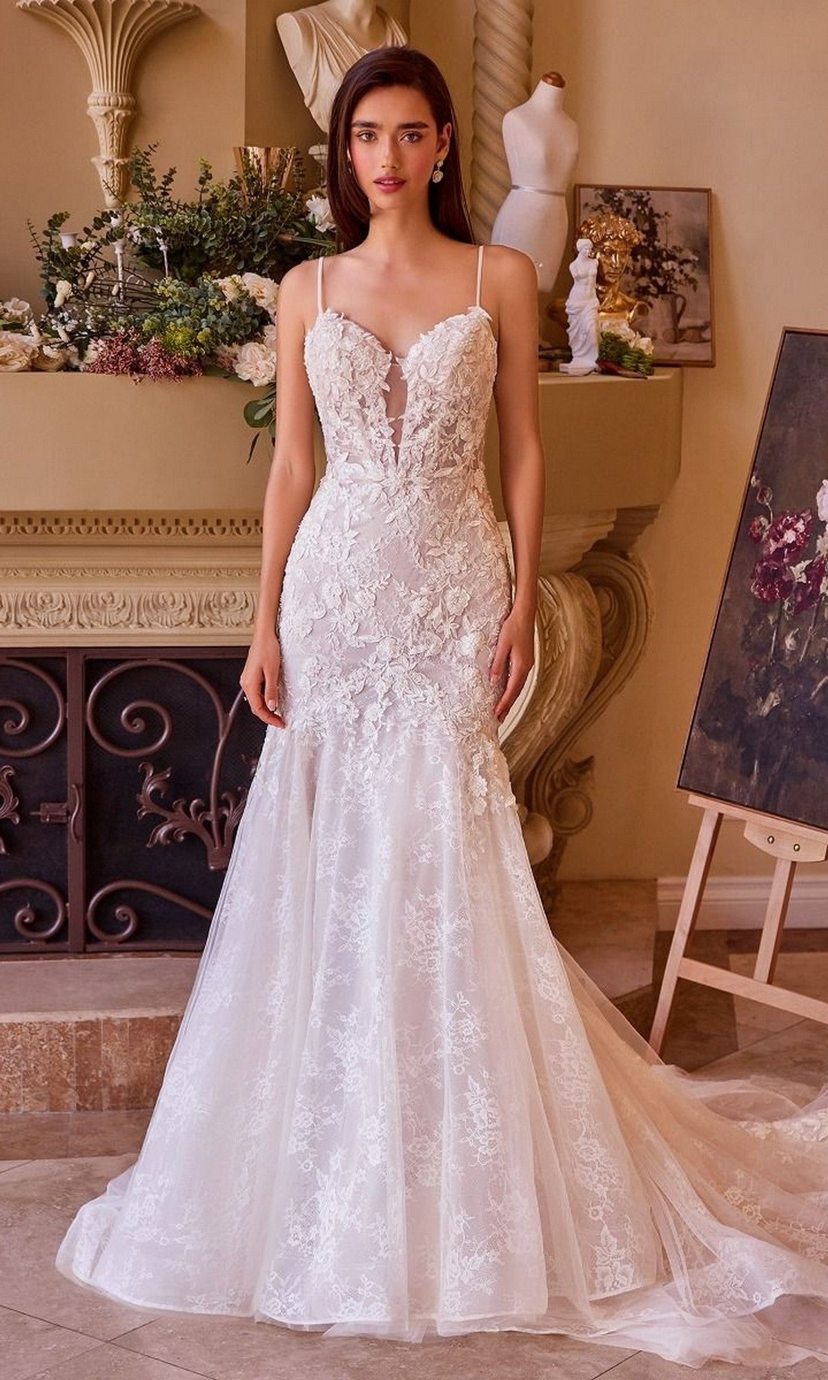 A woman stands indoors in front of a decorated mantelpiece and easel, wearing the Andrea and Leo WL051 long wedding dress— a fitted mermaid gown with thin straps, sheer bodice, and delicate lace applique.